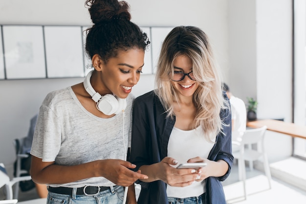 Wunderschöne afrikanische Studentin im Freizeithemd posiert mit Universitätskamerad, der SMS-Nachricht sendet. Attraktive blonde Frau, die als Sekretärin arbeitet und Spaß mit der internationalen Mitarbeiterin im Amt hat.