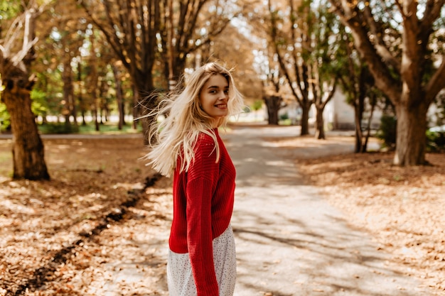 Wunderschöne Blondine, die gute Zeit im schönen Park hat. Hübsches Mädchen, das spielerisch zwischen gelben Blättern lächelt.