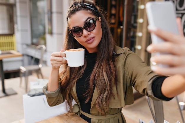 Wunderschöne brünette Frau in der Sonnenbrille, die Kaffee trinkt und sich am Wochenende fotografiert