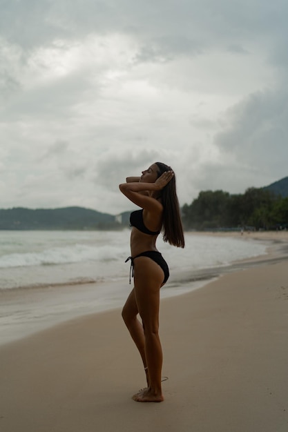 Wunderschöne brünette Frau mit perfekter Figur, die am tropischen Strand posiert und stilvolle schwarze Badebekleidung trägt