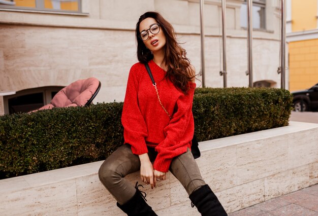 Wunderschöne erfolgreiche brünette Frau, die draußen im trendigen Frühlingsoutfit aufwirft. Modische Stiefel, roter stylischer Pullover. Alte europäische Stadt.