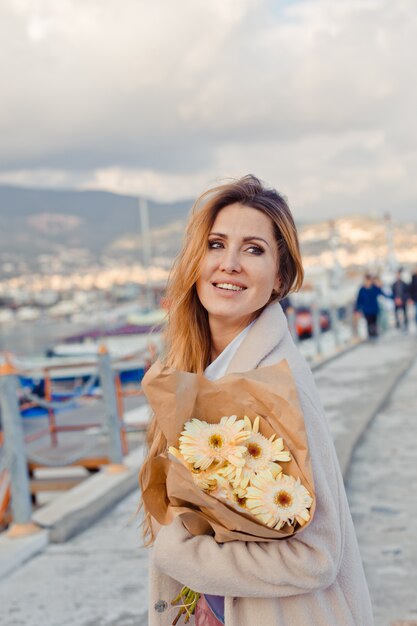 Wunderschöne Frau, die tagsüber am Meer mit Blumen steht und lächelt und liebevoll schaut.