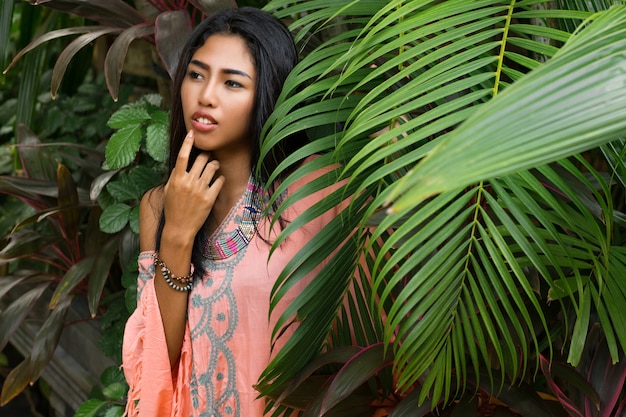Wunderschöne Frau im Boho-Kleid mit grünen Palmblättern. Schöne asiatische Frau in den modischen Sommerkleidern und -zubehör, die im tropischen Naturporträt aufwerfen.
