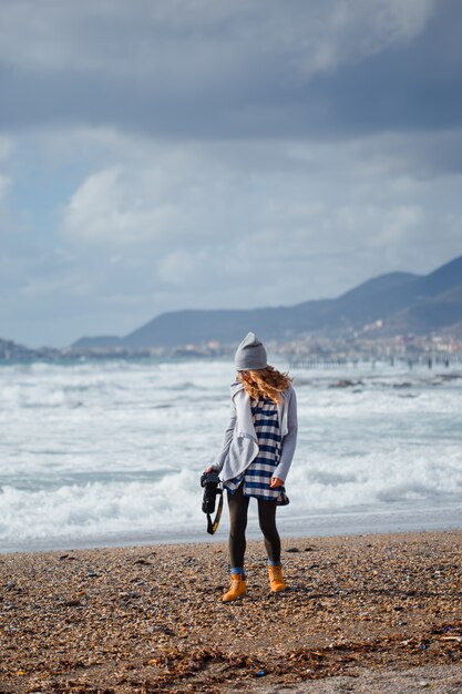 Wunderschöne Frau im grauen Kapuzenpulli und im Hut stehend und hält Kamerastrand während des Tages mit Meer