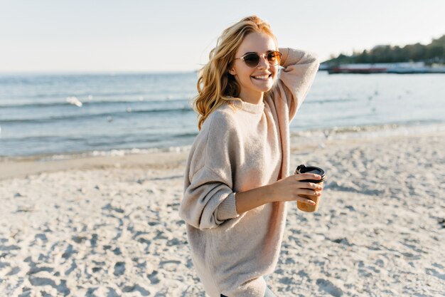 Wunderschöne Frau im Pullover, die an der Seeküste steht. Fashiinable blondhaarige Frau, die Tee nahe Ozean genießt.