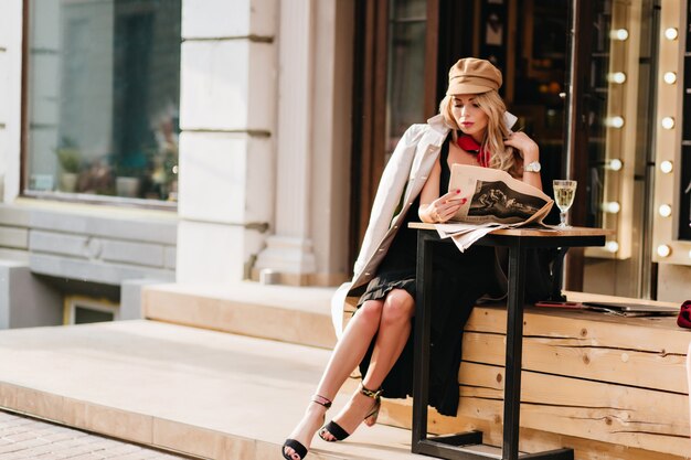 Wunderschöne Frau im schwarzen Kleid, das im Straßencafé ruht und Zeitung liest. Elegantes Mädchen im braunen Mantel und im Hut, die am Tisch mit Glas Champagner und wartendem Freund sitzen.