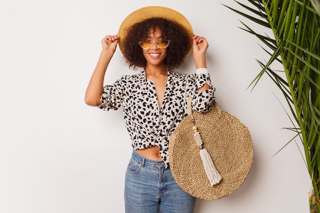 Wunderschöne Frau mit dunkler Haut in Jeans und Strohhut, die im Studio über weißem Hintergrund mit Tasche im Bali-Stil aufwirft. Sopping Stimmung.