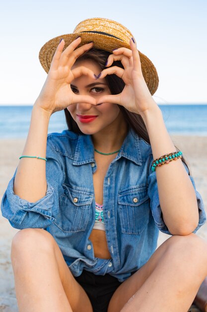 Wunderschöne Frau mit hellbraunem Körper, vollen roten Lippen und l langen Beinen, die auf dem tropischen sonnigen Strand aufwerfen. Tragen von Crop Top, Shorts und Strohhut.