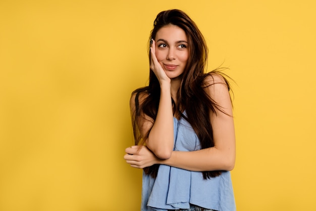 Wunderschöne glückliche Frau im blauen Hemd, das Gesicht mit Hand bedeckt und verträumt in den Rahmen auf gelber Wand schaut.