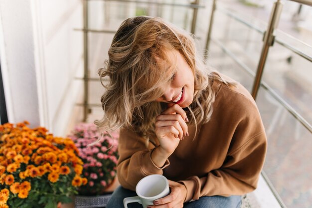 Wunderschöne junge Frau, die Kaffee an der Terrasse trinkt. Frohes kaukasisches Mädchen, das neben orange Blumen mit Lächeln sitzt.