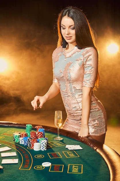 Wunderschöne junge Frau im Abendkleid mit Chips in der Hand, die neben einem Pokertisch mit einem Glas Champagner steht