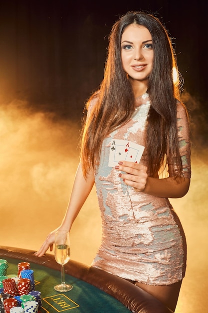 Wunderschöne junge Frau im Abendkleid mit zwei Karten in den Händen, die neben einem Pokertisch mit einem Glas Champagner stehen