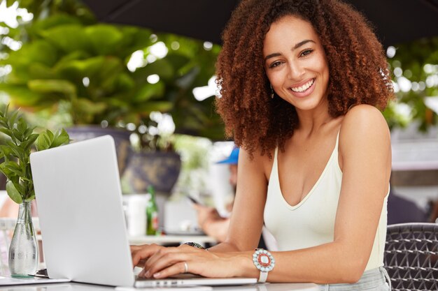 Wunderschöne lächelnde Frau genießt Erholung im Café, hat Videoanruf über tragbaren Laptop, verwendet Anwendung.