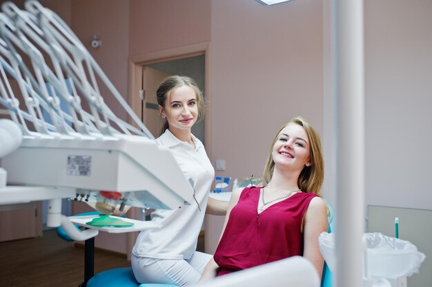 Wunderschöne Zahnärztin posiert und lächelt mit ihrer reizenden Patientin, die auf einem Behandlungsstuhl in rotviolettem Kleid liegt