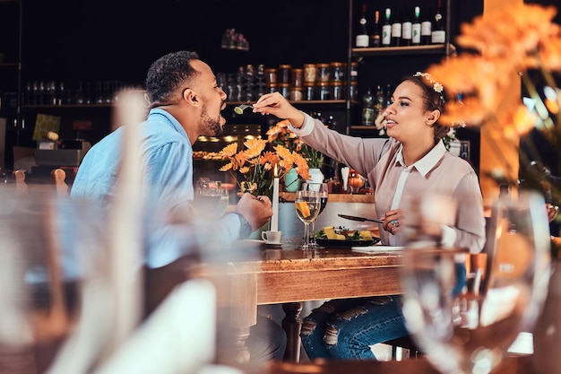 Wunderschönes afroamerikanisches verliebtes Paar, das eine tolle Zeit zusammen bei ihrem Dating hat, ein attraktives Paar, das sich amüsiert, junge Frau, die ihren Mann in einem Restaurant füttert.