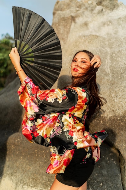 Wunderschönes japanisches Mädchen im stylischen Kimono mit großem Fächer und professionellem Make-up, das über Felsen posiert