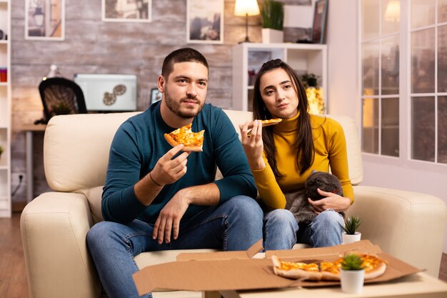 Wunderschönes junges Paar, das Pizza isst, während es im Wohnzimmer auf dem Sofa sitzt?