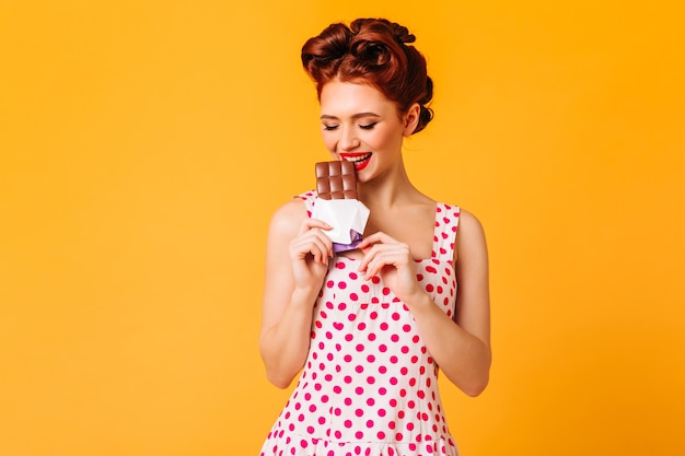 Wunderschönes Mädchen im gepunkteten Kleid, das Schokolade isst. Studioaufnahme der Ingwer-Pinup-Dame, die Nachtisch auf gelbem Raum genießt.