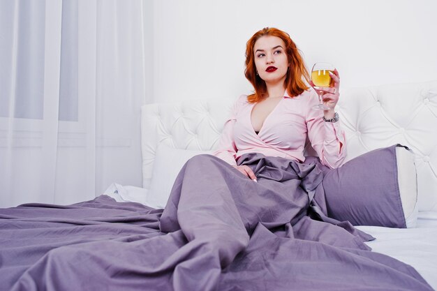 Wunderschönes rothaariges Mädchen in rosa Bluse und rotem Rock mit einem Glas Wein auf dem Bett im Zimmer