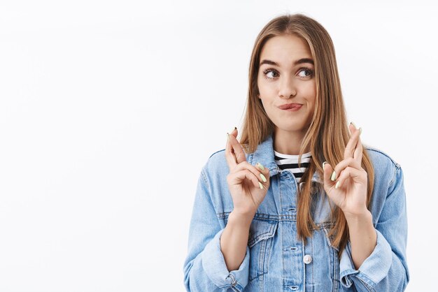 Wunschhaftes süßes Mädchen mit blonden Haaren, das verträumt lächelt und zur Seite schaut und die Daumen für Glück kreuzt, lässt Wunsch, Hoffnung, Traum wahr werden, hat Bestrebungen und Wünsche, steht auf weißem Hintergrund und betet