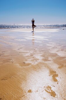 Yoga ausgeglichene haltung für frau in silhouette, die am strand mit blauem meer und wasser steht