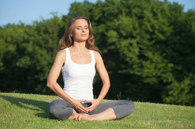 Yogi-Mädchen, das auf dem Gras meditiert Meditation der langhaarigen jungen Frau im weißen Unterhemd auf grünem Hintergrund
