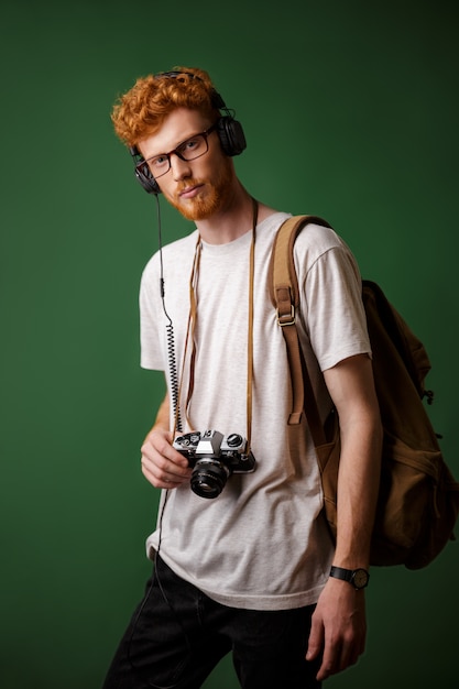 Yong lesekopf bärtiger hipster mit rucksack hält retro-kamera, musik hören,