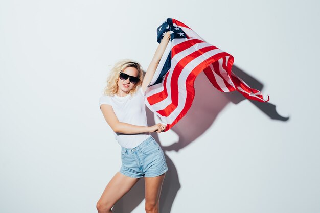 Yung kaukasische blonde Frau in der Sonnenbrille, die USA-Flagge, Unabhängigkeitstag-Feier aufwirft und winkt