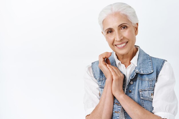Zärtlichkeitsalterung und Kosmetologie-Konzept Moderne, süße und glücklich lächelnde Seniorin mit gekämmten grauen Haaren, die die Hände umklammert und die Kamera anstarrt, freut sich über die Pflege der Haut