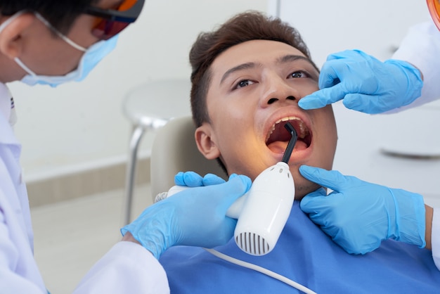 Zahnarzt, der ultraviolettes Licht zum Zahn des asiatischen männlichen Patienten während der Behandlung hält