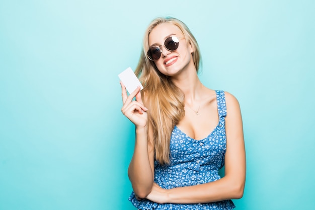 Zahnlächelnde junge Frau in der Sonnenbrille halten Kreditkarte auf blauem Hintergrund.