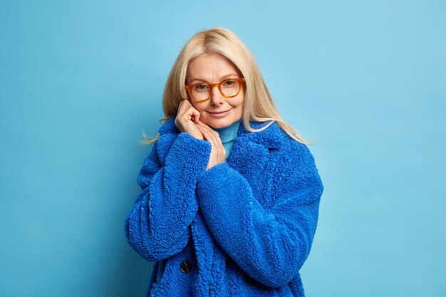 Zarte blonde Europäerin hält Hände in der Nähe von Gesicht sieht angenehm aus hat selbstbewussten Ausdruck trägt Brille modischen Winterpelzmantel.