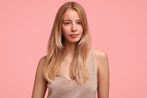 Zarte Frau fühlt sich sanft und schön, trägt lässiges beige T-Shirt, hat glattes helles Haar, isoliert über rosa Wand, sieht ernst aus, zeigt natürliche Schönheit, hat kein Make-up