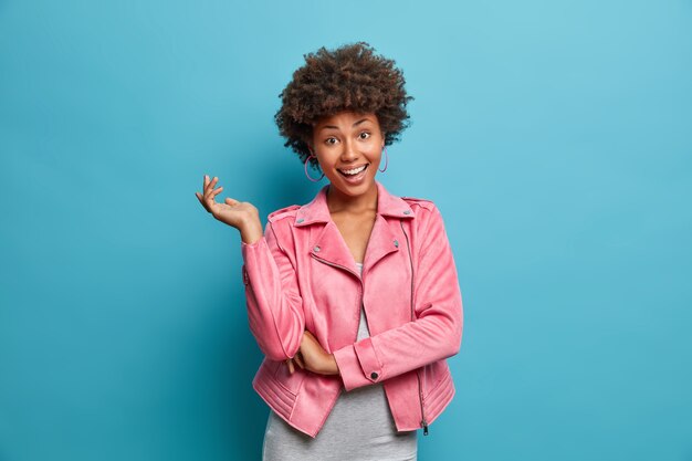 Zarte glückliche junge Afroamerikanerin in rosa Jacke hebt Hand, zeigt perfekte weiße Zähne, freut sich über gute Nachrichten, hat lockiges Haar, posiert