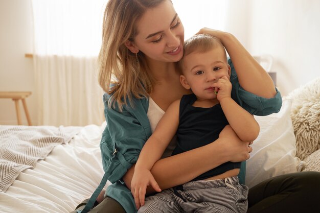 Zarte glückliche junge blonde Mutter sitzt im Schlafzimmer mit charmantem Kleinkindsohn auf ihrem Schoß, schaut ihn mit Liebe und Zuneigung an und streichelt sanft die Haare. Mutter, die mit Babybaby zu Hause verbindet