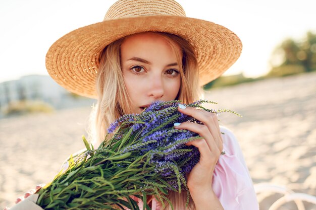 Zarte hübsche Frau im Strohhut, die auf sonnigem Strand nahe Ozean mit Blumenstrauß aufwirft. Nahaufnahme Porträt.