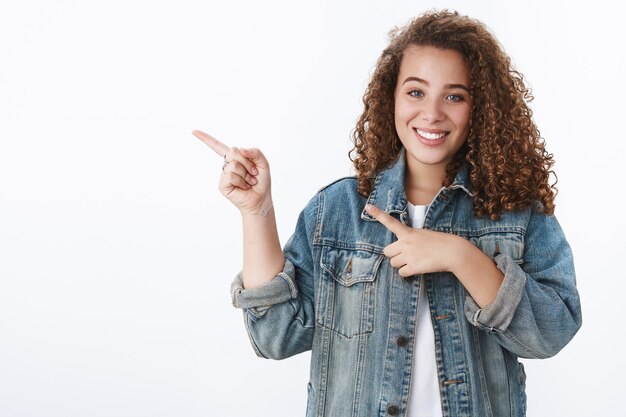 Zarte weibliche junge Studentin lockige Frisur, die mit dem linken Zeigefinger zeigt, lächelt freundlich und fragt, welcher Platz im Wohnheim steht, fröhliche weiße Wand?