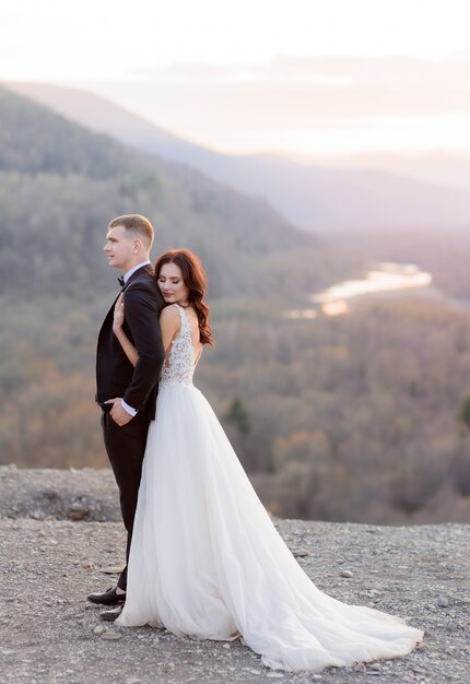 Zartes Hochzeitspaar in der Abenddämmerung auf einem Hügel umarmt sich, gekleidet in luxuriöse Hochzeitskleidung