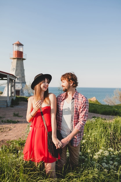 Zartes junges stilvolles Paar verliebt in Landschaft, Indie-Hipster-Bohème-Stil, Wochenendurlaub, Sommeroutfit, rotes Kleid, grünes Gras, Händchen haltend, lächelnd