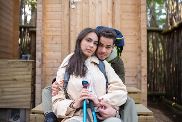 Zartes Paar, das auf der Veranda der Holzhütte sitzt. Frau mit dunklem und bärtigem Mann, der sich umarmt und in die Kamera schaut. Liebe, Zuneigung, Beziehungskonzept