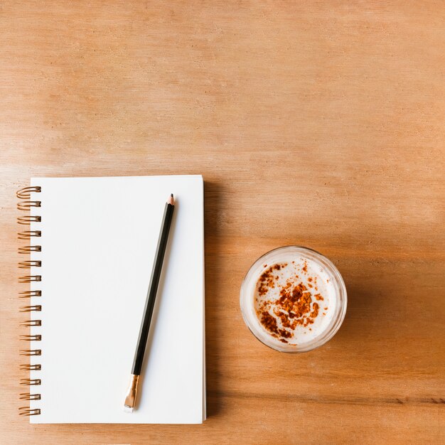 Zeichnen Sie auf geschlossenem weißem gewundenem Notizbuch und Kaffeeglas auf hölzernem strukturiertem Hintergrund an