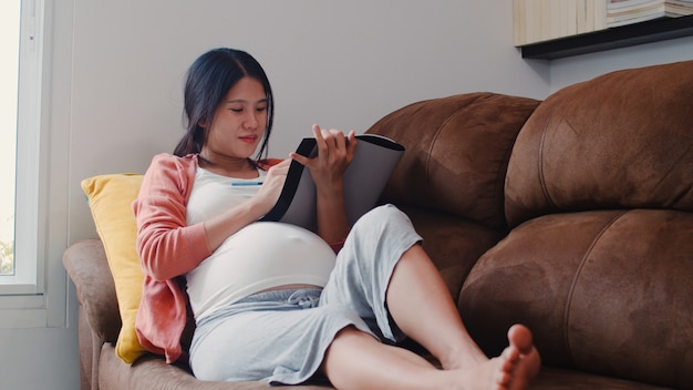 Zeichnungsbaby der schwangeren Frau des jungen Asiaten im Bauch im Notizbuch. Die Mutter, die glücklich sich fühlt, positiv und ruhig lächelnd, während mach s gut das Kind, das zu Hause auf Sofa im Wohnzimmer liegt.