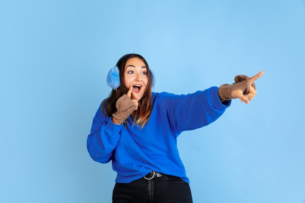 Zeigen. porträt der kaukasischen frau auf blauem studiohintergrund. schönes weibliches modell in warmer kleidung. konzept der menschlichen emotionen, gesichtsausdruck, verkauf, anzeige. winterstimmung, weihnachtszeit, feiertage.