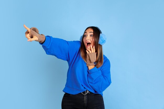 Zeigen. Porträt der kaukasischen Frau auf blauem Studiohintergrund. Schönes weibliches Modell in warmer Kleidung. Konzept der menschlichen Emotionen, Gesichtsausdruck, Verkauf, Anzeige. Winterstimmung, Weihnachtszeit, Feiertage.