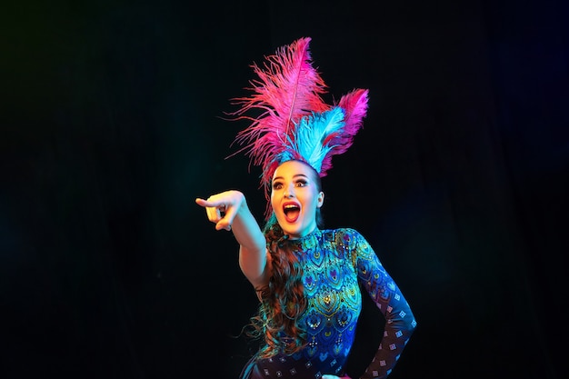 Zeigen. Schöne junge Frau im Karneval, stilvolles Maskenkostüm mit Federn auf schwarzem Hintergrund im Neonlicht. Exemplar für Anzeige. Feiertagsfeier, Tanzen, Mode. Festliche Zeit, Party.