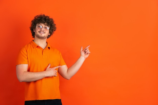 Zeigen, wählen. Das monochrome Portrait des kaukasischen jungen Mannes an der orangefarbenen Wand. Schönes männliches lockiges Modell im lässigen Stil. Konzept der menschlichen Emotionen, Gesichtsausdruck, Verkauf, Anzeige.