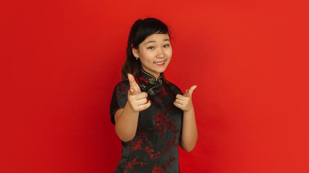 Zeigen, wählen, lächeln. frohes chinesisches neues jahr 2020. porträt des asiatischen jungen mädchens auf rotem hintergrund. weibliches modell in traditioneller kleidung sieht glücklich aus. feier, menschliche gefühle. copyspace.
