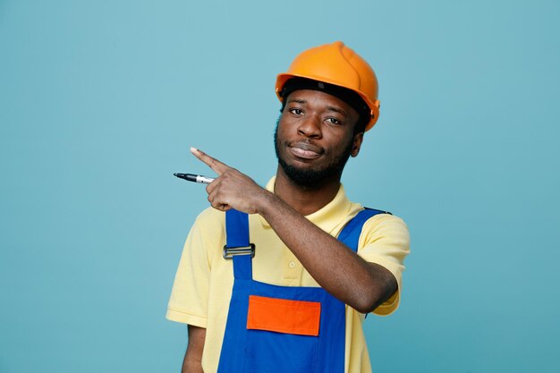 Zeigt auf die Seite des jungen afroamerikanischen Baumeisters in Uniform, der einen Bleistift isoliert auf blauem Hintergrund hält