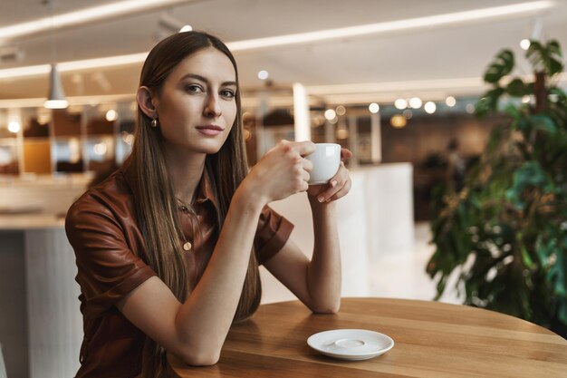 Zeit, frisch gebrühten Kaffee zu genießen Attraktive entspannte junge weibliche Frau in braunem Kleid, die allein im Café sitzt und an einem friedlichen, gemütlichen Ort Cappuccino mit lächelnder Kamera hält