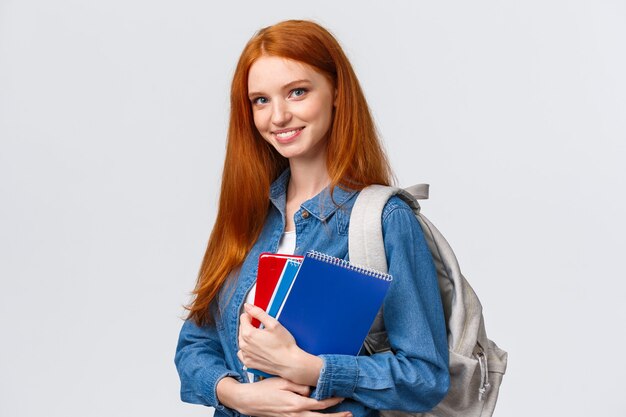 Zeit zur Schule. Schöne fröhliche moderne rothaarige Frau mit Rucksack, die Notebooks hält, die auf das College gehen, amüsiert lächeln, nach der Pause zurück in die Klasse gehen, weißer Hintergrund stehen